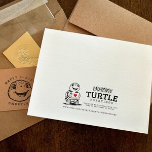 May include: A white card with a black and white illustration of a turtle holding a red heart with the text "Happy Turtle Greetings" printed on it. The card is designed and printed with love in the heart of Texas. The text "www.etsy.com/shop/HappyTurtleGreetings" is printed at the bottom of the card.