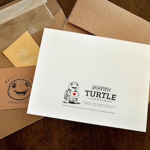 May include: A collection of stationery items, including a greeting card with a turtle illustration holding a heart, a business card, and envelopes. The card features the text "Happy Turtle Greetings."