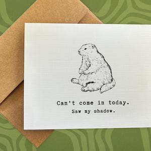 May include: A white card with a black and white illustration of a groundhog. The card reads "Can't come in today. Saw my shadow."