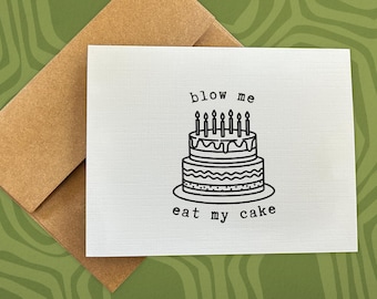 May include: A white card with black text that reads "blow me eat my cake". A black and white illustration of a birthday cake with candles is in the center of the card.