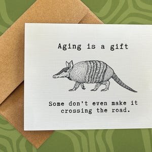 May include: A white card with black text that reads "Aging is a gift. Some don't even make it crossing the road." A black and white illustration of an armadillo is in the center of the card.