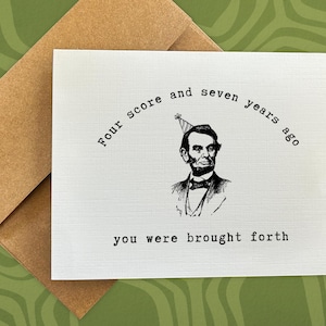 May include: A white card with black text that reads "Four score and seven years ago you were brought forth". A black and white illustration of Abraham Lincoln wearing a party hat is above the text.