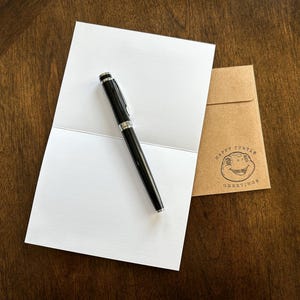 May include: A blank white card with a black pen on top of it. The card is folded in half. A brown envelope with the text "Happy Turtle Greetings" is in the background.