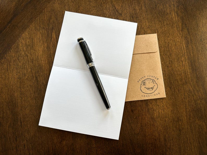 May include: A blank white card with a black pen on top of it. The card is folded in half. A brown envelope with the text "Happy Turtle Greetings" is in the background.