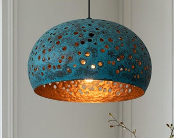 Puede incluir: Una lámpara colgante turquesa y cobre. La pantalla en forma de cúpula tiene un diseño texturizado y perforado, con un interior de cobre. La luz cuelga de un cable negro, iluminando el espacio inferior. La lámpara es un elemento decorativo.