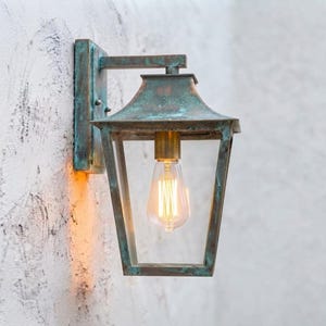 May include: A wall-mounted lantern light fixture featuring a weathered verdigris copper finish with a rectangular frame and a sloped roof. The fixture holds a single exposed Edison-style light bulb with a visible filament, emitting a warm glow.