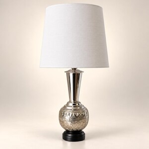 May include: A table lamp featuring a white tapered fabric shade atop a polished silver-toned metal base. The base consists of a smooth, slender neck rising from a rounded, textured body with intricate embossed patterns, all set on a black circular stand.