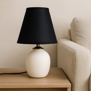 May include: A small, white ceramic table lamp with a black lampshade. The lamp has a rounded base and a brass-colored neck. The lampshade is cone-shaped. The lamp is on a wooden table next to a beige sofa.
