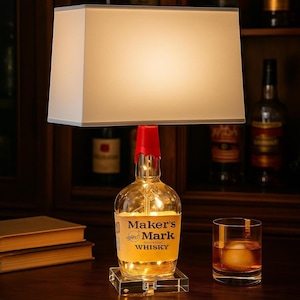 May include: A unique lamp made from a Maker's Mark whiskey bottle, featuring a red wax seal and a rectangular lampshade. The bottle base is illuminated with small lights, and the lamp sits on a clear, square base. A glass of whiskey with an ice sphere is on the right.