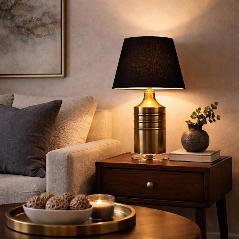 Op de afbeelding: Een goudkleurige tafellamp met een zwarte lampenkap. De lamp heeft een cilindrische voet met horizontale ribbels en een heldere glazen voet. De lamp staat op een donkerhouten nachtkastje naast een kleine vaas en een stapel boeken.