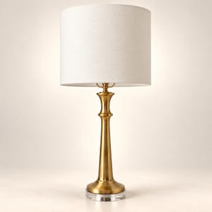 May include: A table lamp featuring a tall, slender, brushed brass metal base with a flared bottom and a clear circular glass pedestal. The lamp is topped with a cylindrical, off-white fabric drum shade that provides a neutral, clean aesthetic.