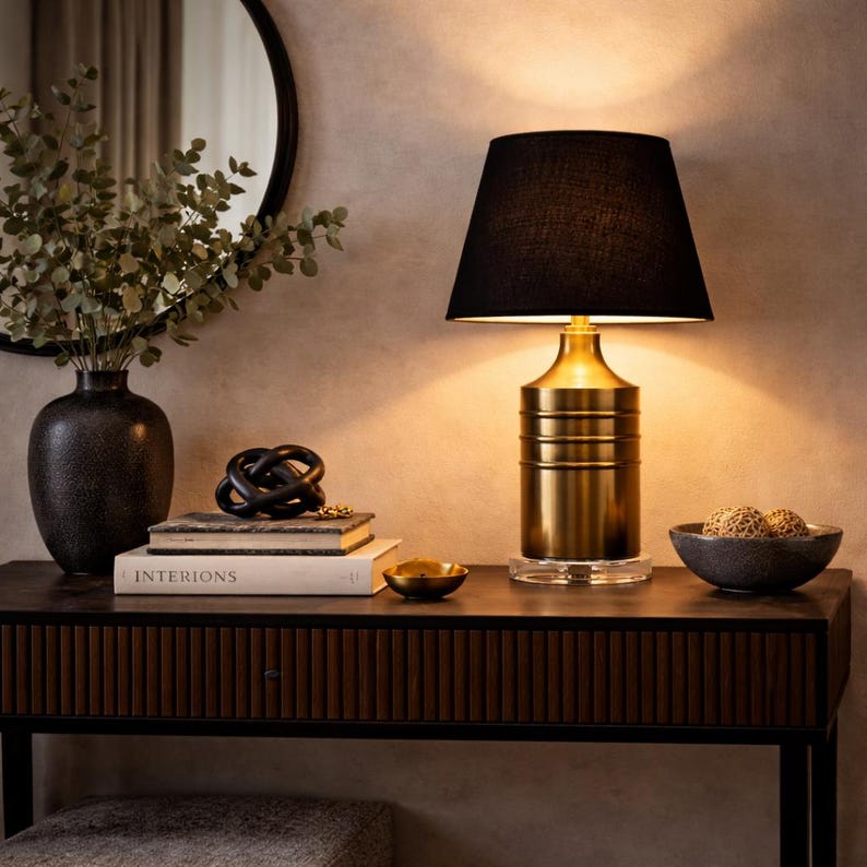 Op de afbeelding: Een goudkleurige tafellamp met een zwarte lampenkap staat op een donkere houten tafel. De lamp heeft een cilindrische voet en een heldere, vierkante basis. Andere decoraties zijn een zwarte vaas met groen, boeken en een kleine kom.