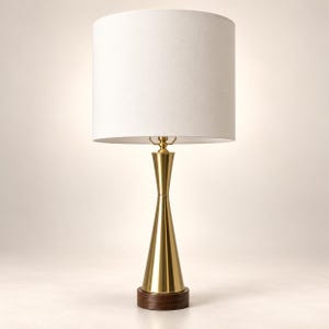 May include: A table lamp featuring a cylindrical white fabric shade atop a slender, polished gold-tone metal base with a tapered hourglass silhouette. The lamp stands on a circular dark brown wooden base, providing a stable foundation for the fixture.
