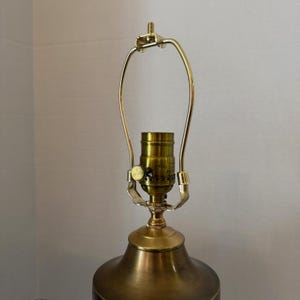 Op de afbeelding: Close-up van een messingkleurige lampvoet en hardware. De lamp heeft een gebogen, goudkleurige harp en een messingkleurige fitting. De voet heeft een afgeronde vorm met een donkere, mogelijk geschilderde afwerking. De lamp is ontworpen voor een lampenkap.