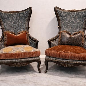 May include: Two antique armchairs with high backs. The chairs have a dark wood frame and are upholstered in a patterned blue fabric. Each chair has a decorative orange pillow and a fringed seat cushion. The chairs are placed on a light-colored floor.