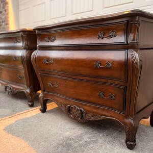 May include: A pair of dark brown wooden chests of drawers featuring three horizontal drawers each. The furniture pieces have curved fronts, ornate carved detailing on the legs and apron, and dark metal handles on every drawer front.