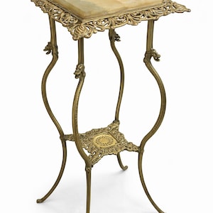 May include: An antique brass plant stand with a square marble top. The stand features ornate, scrolling details and curved legs. The stand is approximately 30 inches tall.