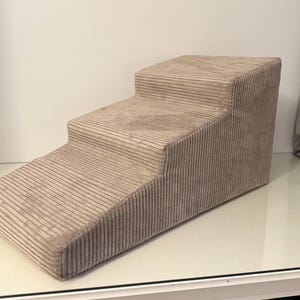 Può includere: Un gradino per animali domestici in velluto a coste beige con due gradini e una rampa. I gradini sono progettati per aiutare cani o gatti di piccola taglia a raggiungere superfici più alte. Il tessuto ha una trama a coste.