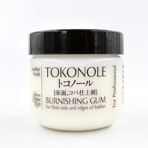 Op de afbeelding: Een witte pot met een zwarte deksel met het label "Tokonole" in zwarte tekst. De pot bevat polijstgom voor leer, en het etiket bevat ook de Japanse tekst "トコノール" en de zin "[床面、コバ仕上剤] BURNISHING GUM for flesh side and edges of leather".