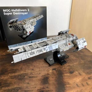 Puede incluir: Un modelo gris y blanco de una nave espacial Super Destroyer, exhibido sobre un soporte negro. El modelo está detallado con varias formas y texturas. La caja detrás del modelo dice "MOC-Helldivers 2 Super Destroyer."