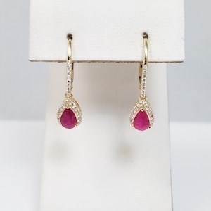 May include: A pair of gold hoop earrings with a teardrop-shaped ruby gemstone pendant. The ruby is surrounded by a halo of small, clear gemstones. The earrings are displayed on a white stand.
