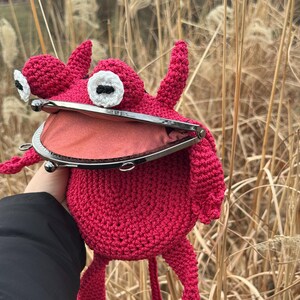 May include: A crocheted red monster coin purse with a large open mouth and two large white eyes. The purse has a metal clasp and a long tail.
