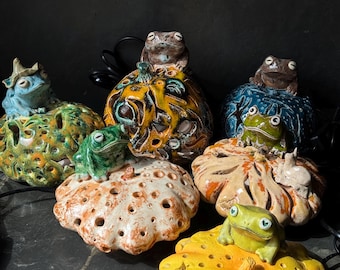May include: A collection of six ceramic pumpkin-shaped lanterns, each topped with a sculpted frog figure. The pumpkins feature various glaze colors including yellow, orange, green, and blue, with cut-out patterns and black electrical cords attached.