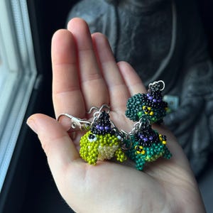Può includere: Portachiavi a forma di rana fatti a mano con perline nei toni del verde, nero e viola. Ogni rana indossa un piccolo cappello da strega e ha un fermaglio argentato. I portachiavi sono dettagliati con piccole perline, creando un aspetto strutturato.