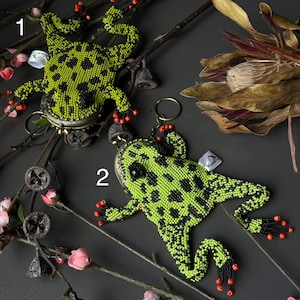 May include: Two beaded frog-shaped keychains. The frogs are lime green with black spots and red accents on their feet. They are set against a dark gray background with floral and twig accents.