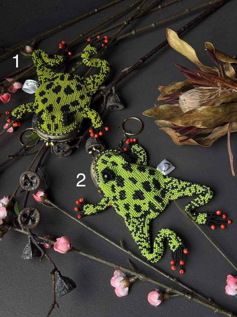 May include: Two beaded frog-shaped keychains. The frogs are lime green with black spots and red accents on their feet. They are set against a dark gray background with floral and twig accents.