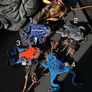 May include: Four beaded frog-shaped coin purses in blue, orange, and gray. The frogs have detailed beadwork, with some featuring white stripes or black spots. Each purse has a metal clasp and a small tag.