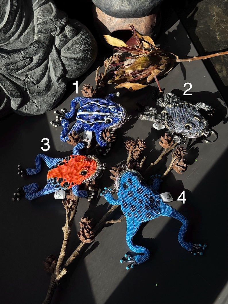 May include: Four beaded frog-shaped coin purses in blue, orange, and gray. The frogs have detailed beadwork, with some featuring white stripes or black spots. Each purse has a metal clasp and a small tag.