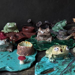 Puede incluir: Una colección de figuras de ranas de cerámica sobre bases de hojas de nenúfar. Las ranas están esmaltadas en tonos verdes y marrones, algunas con pequeños detalles de setas, formas de troncos y portavelas integrados en las plataformas.