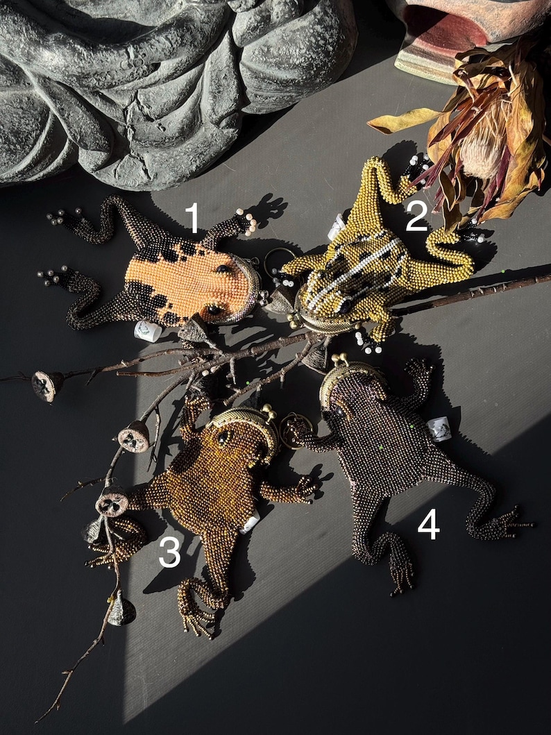 May include: Four beaded frog-shaped coin purses in various colors. One is orange and black, another is yellow and white, and the other two are shades of brown. Each has a clasp and is posed on a dark surface with a branch.