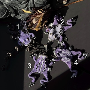 May include: Four beaded frog-shaped ornaments in varying shades of purple, with black and white accents. Each frog has a silver-toned clasp. The frogs are arranged on a dark surface, with a few dried plants in the background.