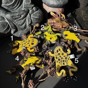 May include: Five beaded frog ornaments in yellow and black, arranged on a dark surface with dried branches and pinecones. The frogs vary in size and pose, showcasing intricate beadwork details.
