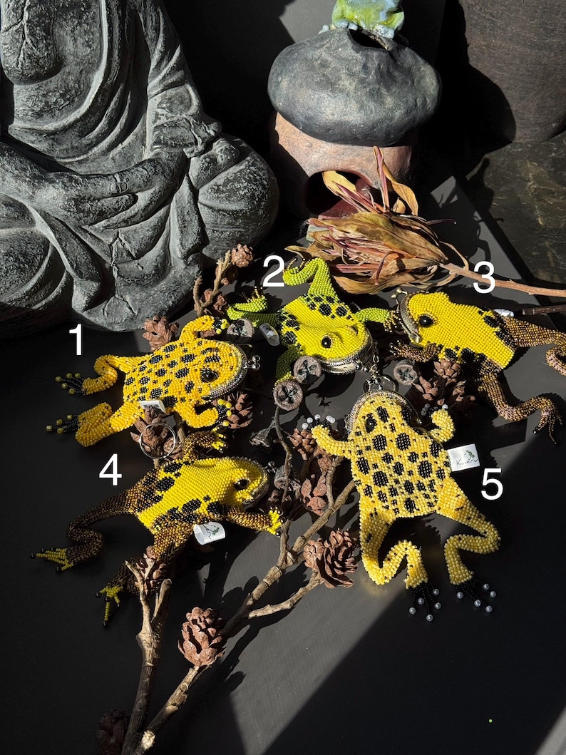 May include: Five beaded frog ornaments in yellow and black, arranged on a dark surface with dried branches and pinecones. The frogs vary in size and pose, showcasing intricate beadwork details.