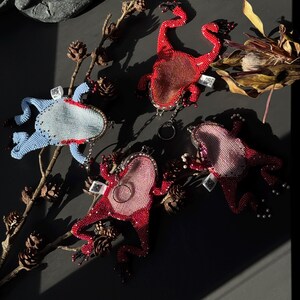 May include: A collection of beaded frog-shaped keychains in various colors, including red, pink, and blue. The keychains are embellished with small beads and feature intricate details. The frogs are arranged on a dark surface with natural elements.