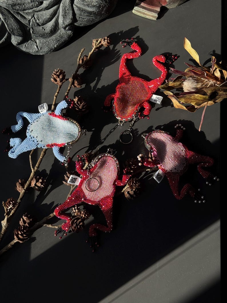 May include: A collection of beaded frog-shaped keychains in various colors, including red, pink, and blue. The keychains are embellished with small beads and feature intricate details. The frogs are arranged on a dark surface with natural elements.