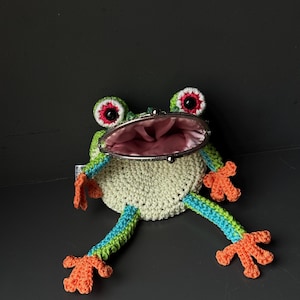 Könnte beinhalten: Eine handgefertigte, gehäkelte Frosch-Geldbörse. Der Frosch hat einen hellgrünen Körper, große rot-weiße Augen und orangefarbene Füße. Die Geldbörse hat einen silbernen Verschluss und ein rosa Innenfutter mit blau-grün gestreiften Beinen.
