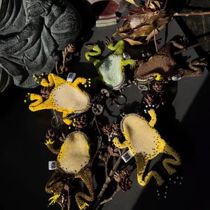 May include: A collection of beaded frog-shaped keychains in various colors, including yellow, green, and brown. The keychains are arranged on a dark surface with dried foliage and pinecones. Each frog has a unique design.