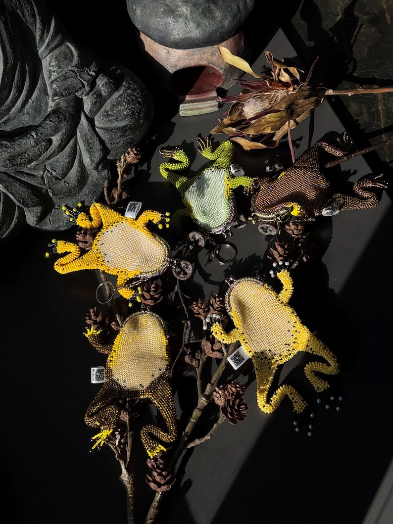 May include: A collection of beaded frog-shaped keychains in various colors, including yellow, green, and brown. The keychains are arranged on a dark surface with dried foliage and pinecones. Each frog has a unique design.