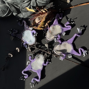 May include: Four beaded frog keychains in a vibrant purple hue, each with a silver-colored belly. The frogs are arranged on a dark surface with dried foliage and a stone sculpture in the background. The keychains have white and black beaded accents.