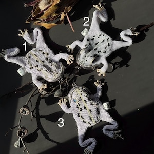 May include: Three beaded frog-shaped coin purses. Each frog is white with black and yellow spots, and has a silver-toned clasp. The frogs are arranged on a dark surface with dried plant elements.