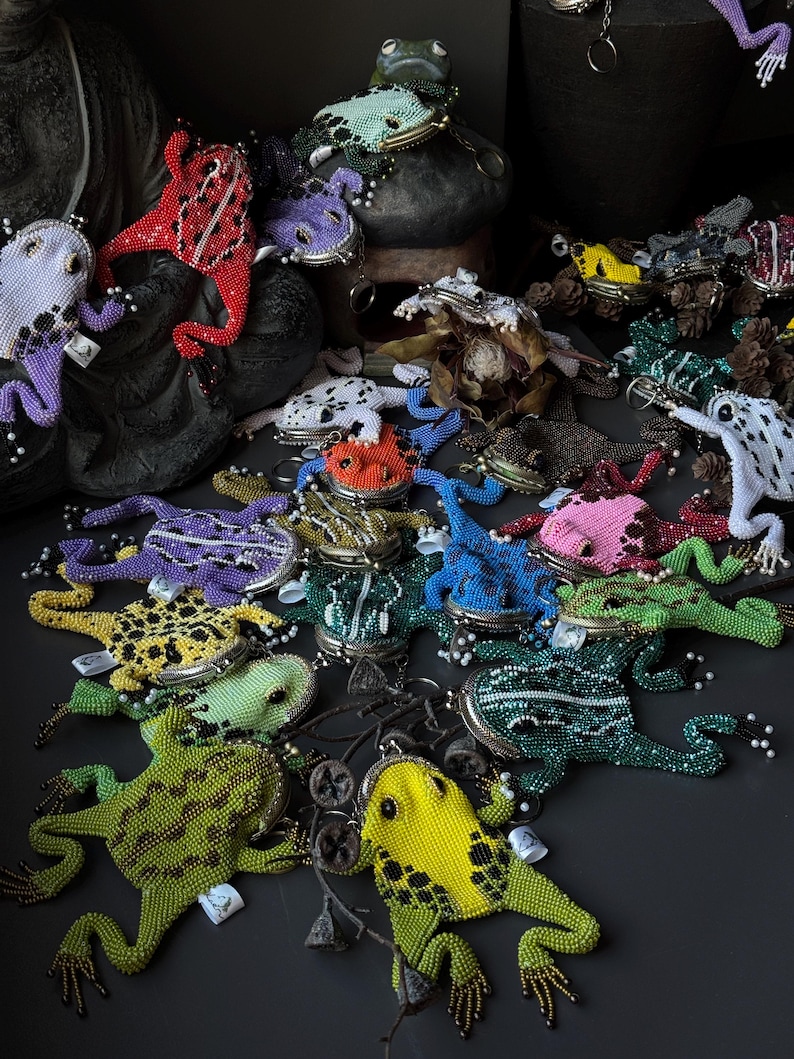 May include: A collection of beaded frog-shaped coin purses in various colors, including green, yellow, red, purple, and blue. Each purse features intricate beadwork and a metal clasp. Some purses are arranged on a dark surface, while others are perched on a stone sculpture.