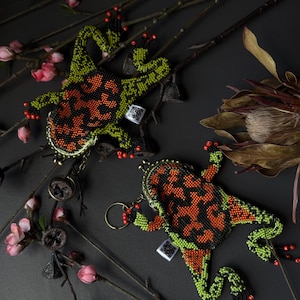 May include: Two beaded frog-shaped coin purses. The frogs have a black, orange, and green pattern. The purses have a metal clasp and a keychain ring. The background includes branches, pink flowers, and a dark surface.
