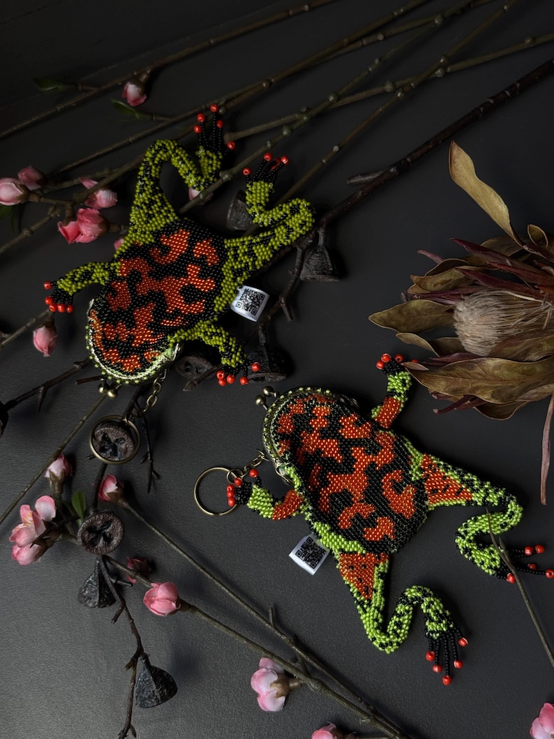 May include: Two beaded frog-shaped coin purses. The frogs have a black, orange, and green pattern. The purses have a metal clasp and a keychain ring. The background includes branches, pink flowers, and a dark surface.