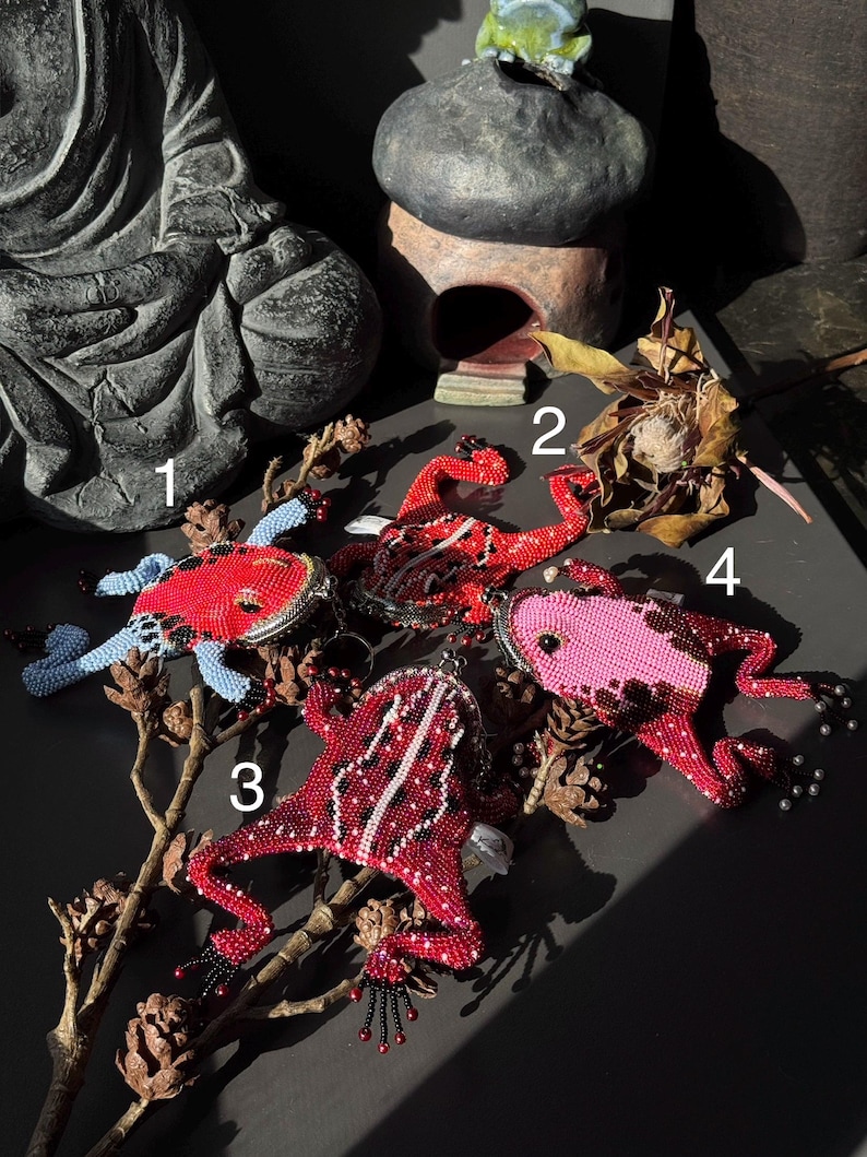 May include: Four beaded frog ornaments in red, pink, and blue hues. Each frog has intricate beadwork, with details like beaded eyes and feet. The frogs are arranged on a dark surface with dried branches and pinecones.