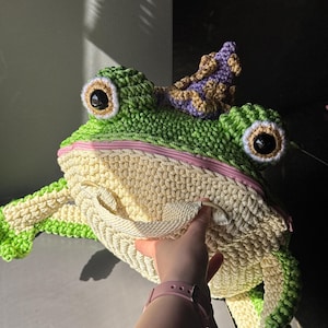 May include: A large, handmade crochet frog bag in shades of green and cream. The frog has large, round eyes, a pink zipper, and a small crown. The bag is being opened by a hand.