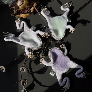 May include: Three beaded frog-shaped accessories with silver-tone accents. The frogs have white beaded bodies and light green or purple centers. Each frog has a small crown and tiny bells. The accessories are arranged on a dark surface.